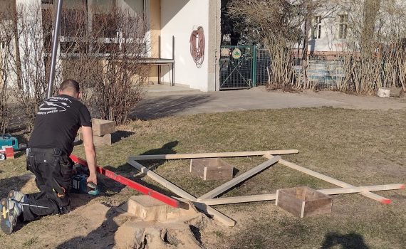 Kita Müggelzwerge, Beteiligungsprojekt Tippi, Handwerker mit Saege und Wasserwaage auf der Wiese baut einen Rahmen für eine Tippi-Zelt