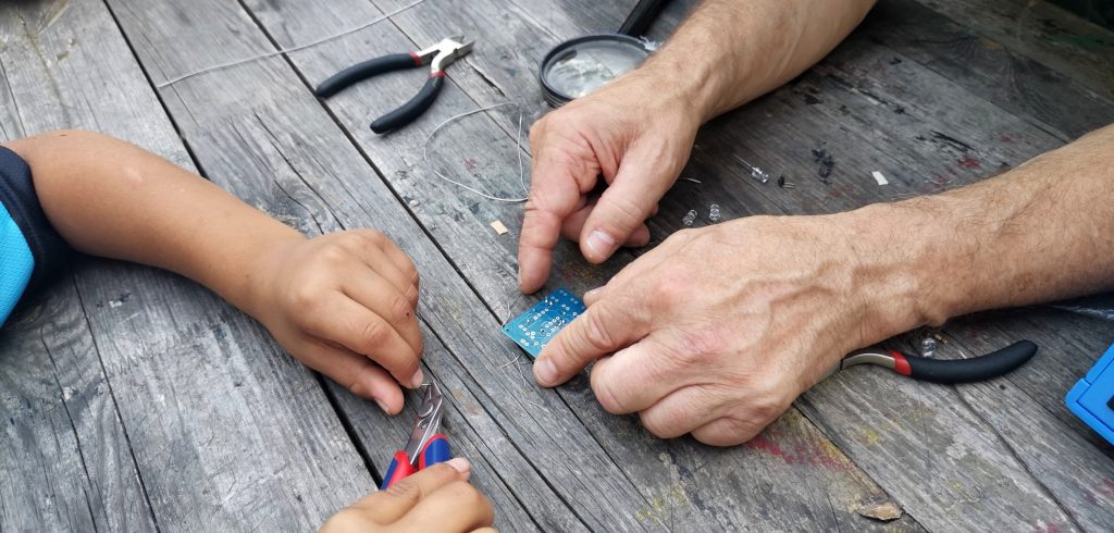 FAZ Fennpfuhl Elektronikbasteln, Erwachsenenhände zeigen Kinderhände eine Schaltplatte